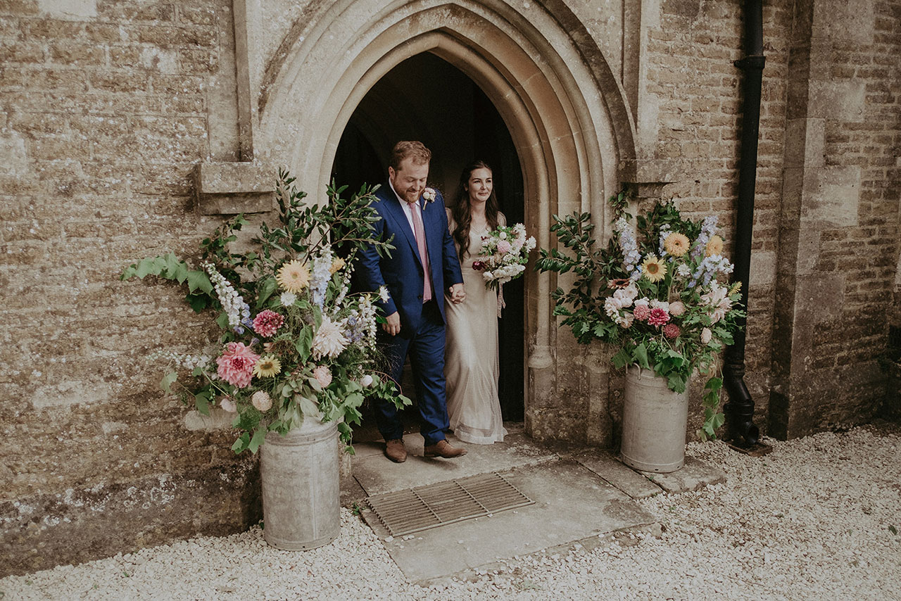 Bride and Groom leaving church with bridal bouquet through church porch with flower buckets either side